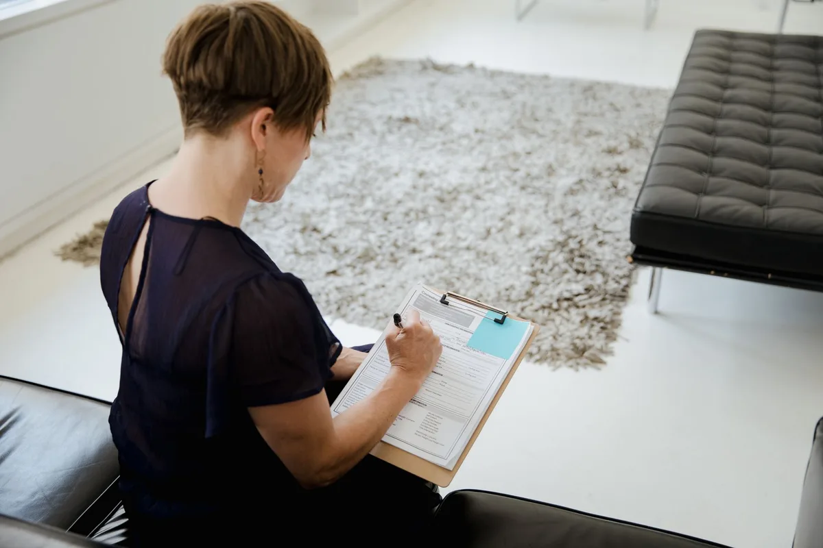 A woman sitting indoors writing on a clipboard, focused on completing an assessment