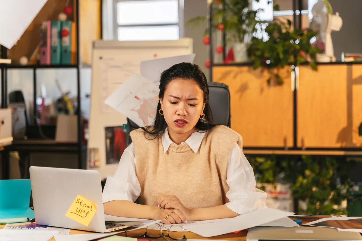 A frustrated creator sitting at a cluttered desk, staring at a laptop with a &ldquo;Start Up&rdquo; sticky note among scattered papers